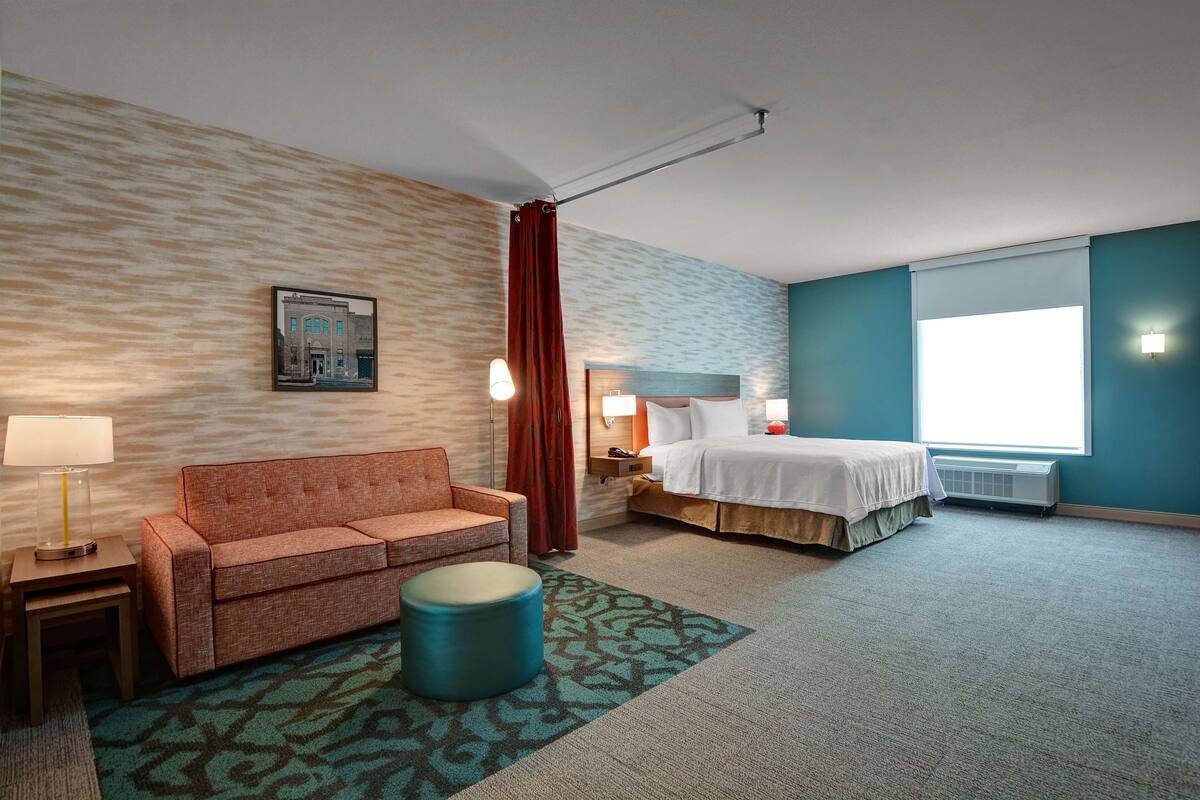 Une chambre d’hôtel comprenant un canapé, un pouf rond, un lit, une table de chevet, une lampe et une fenêtre avec des stores.