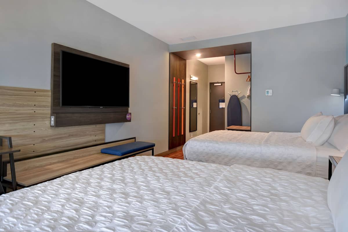 A hotel room with two beds, a TV mounted on the wall, a bench, and a mirror.