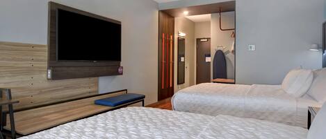 A hotel room with two beds, a TV mounted on the wall, a bench, and a mirror.