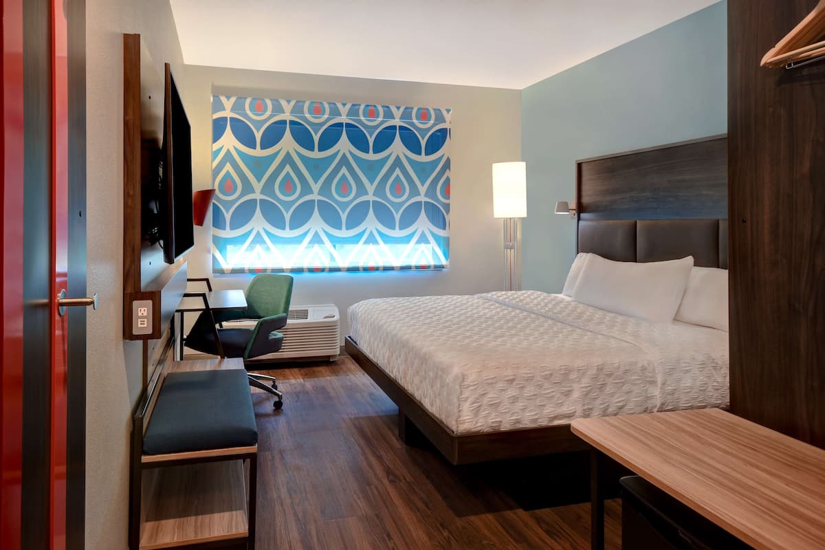 A hotel room with a bed, a desk, a chair, a TV, and a window with a patterned curtain.