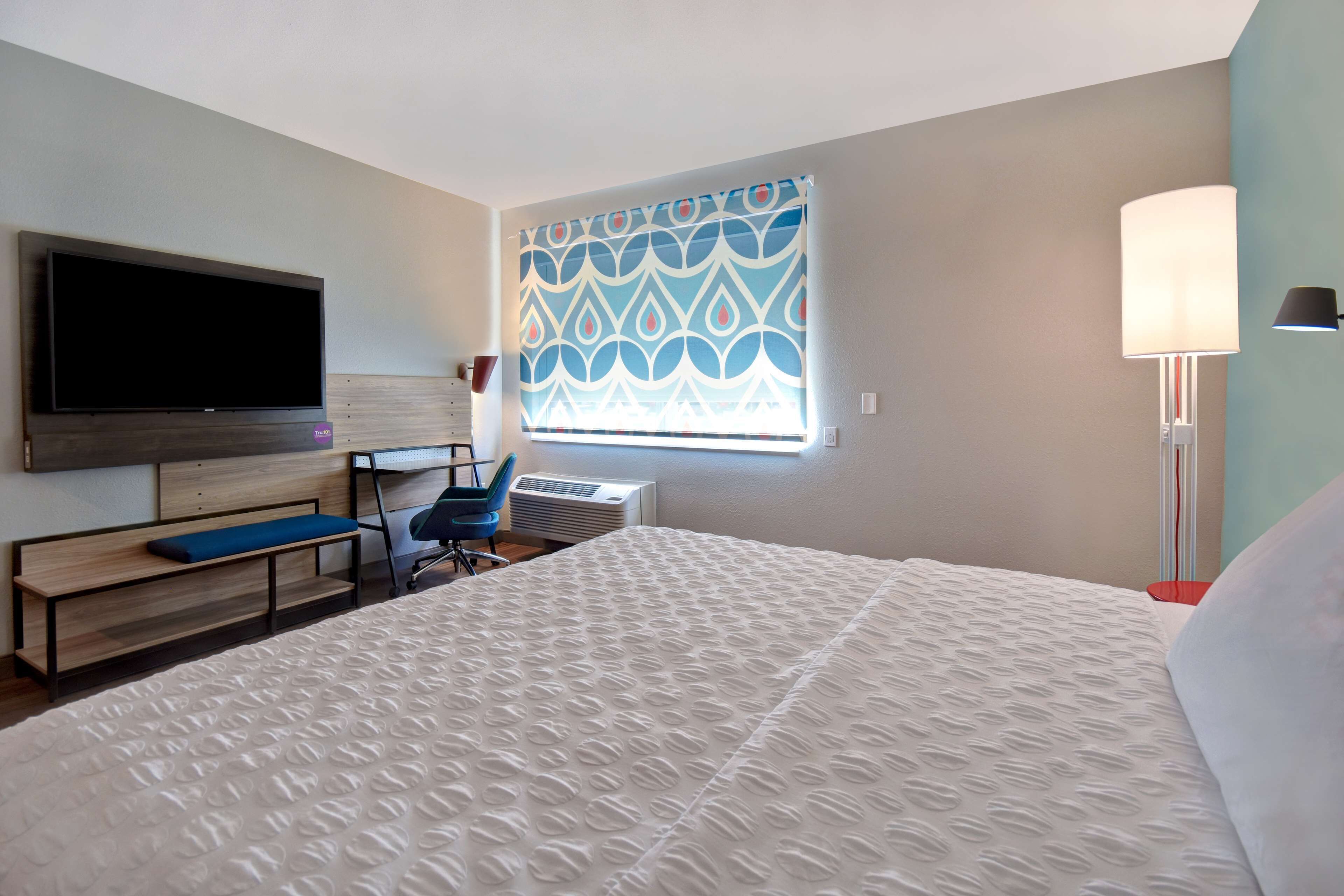 A hotel room with a large bed, a flat-screen TV, a desk with a chair, a window with patterned curtains, and a standing lamp.