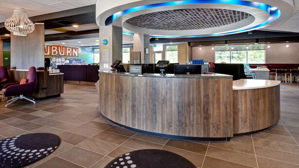 A hotel reception area with a curved reception desk, a seating area with purple chairs, and a large window providing a view of the outdoors.