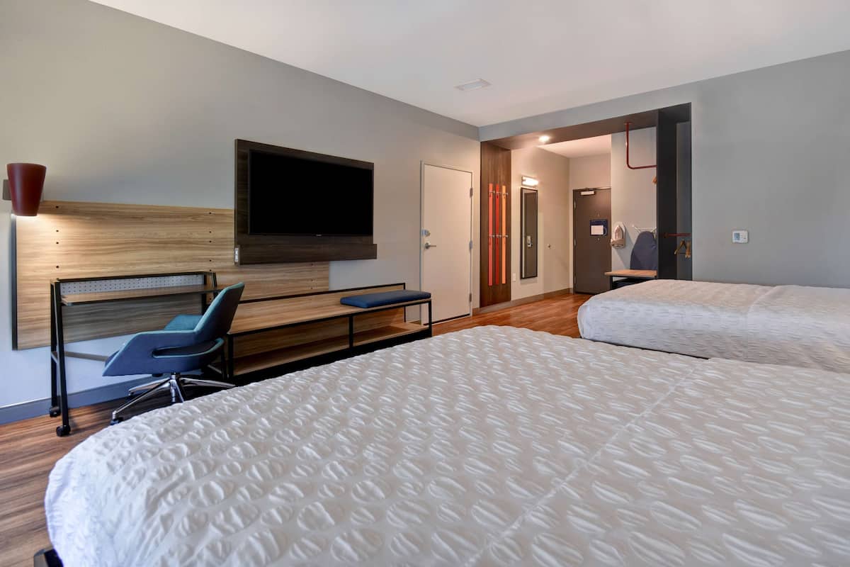 A hotel room with two beds, a desk, a chair, a TV mounted on the wall, and a door leading to a bathroom.