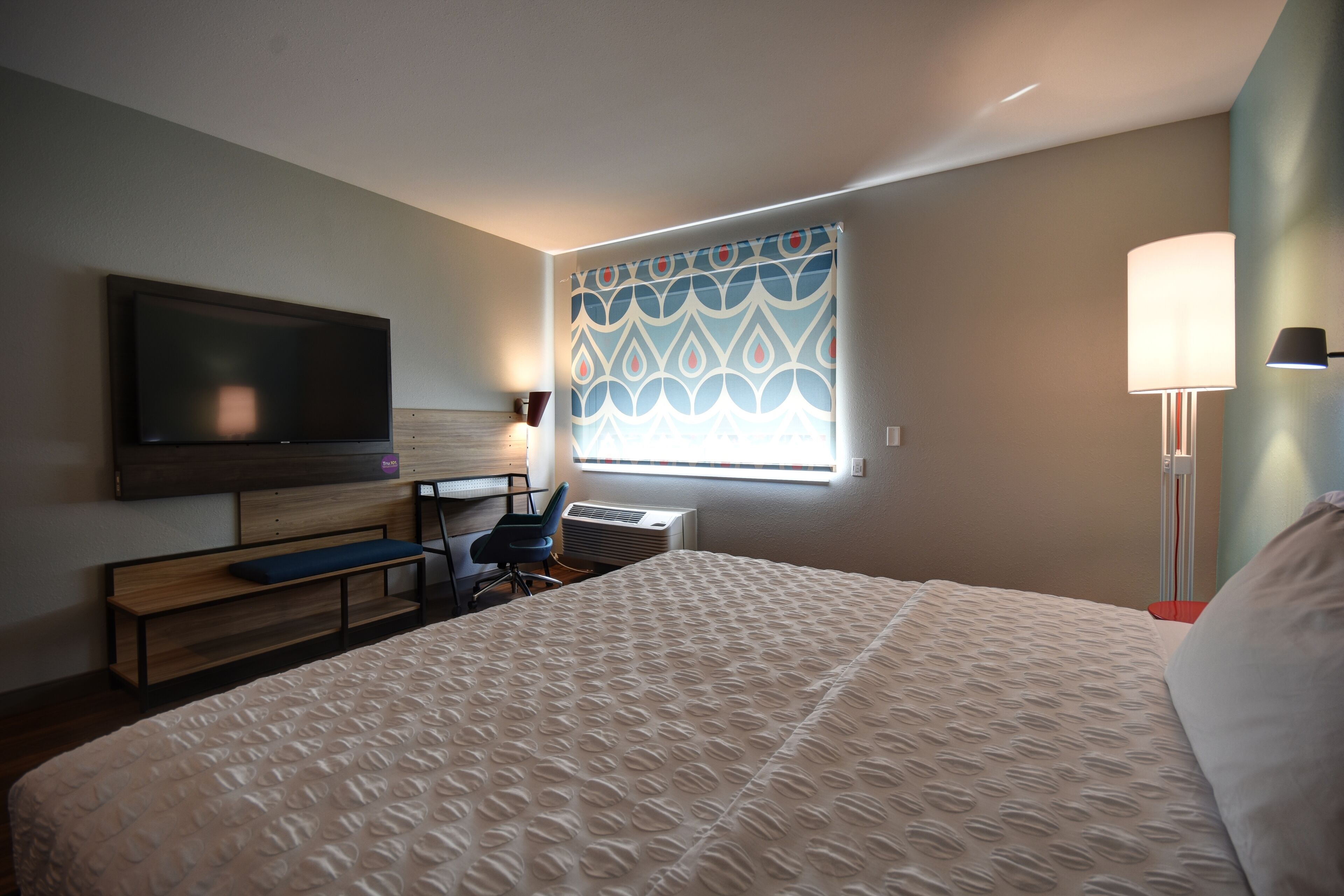 A hotel room with a large bed, a desk with a chair, a flat-screen TV mounted on the wall, and a window with patterned curtains.