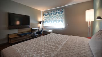 A hotel room with a large bed, a desk with a chair, a flat-screen TV mounted on the wall, and a window with patterned curtains.