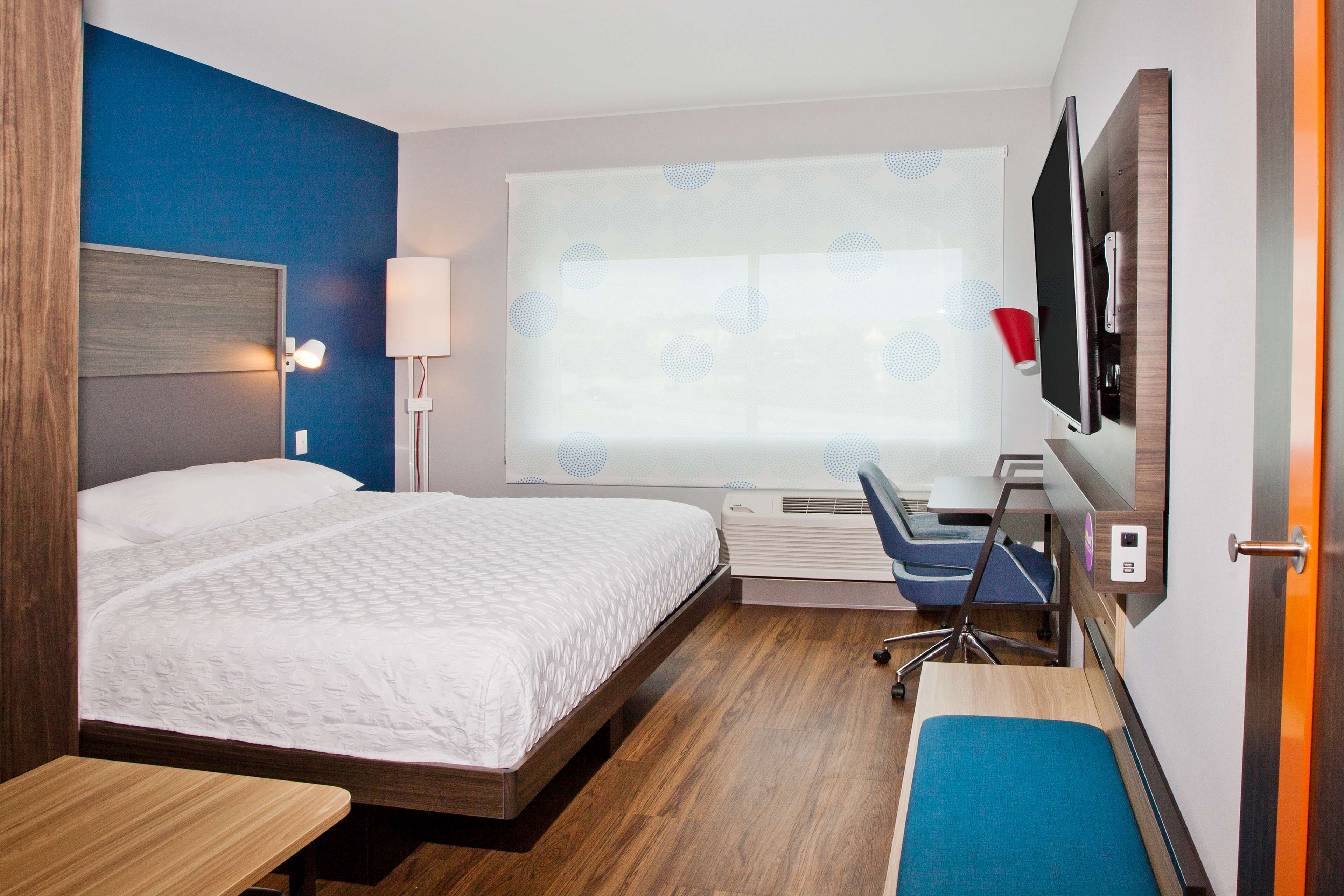 A hotel room with a bed, desk, chair, TV, and window with blinds.