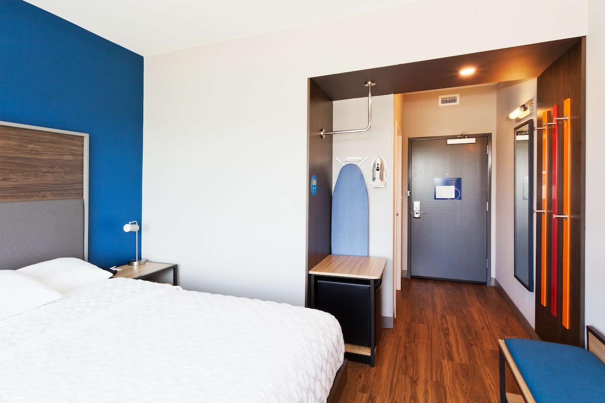 Ein Hotelzimmer mit Bett, Nachttisch und Spiegel. Ein Flur mit einer Tür zu einem Badezimmer.