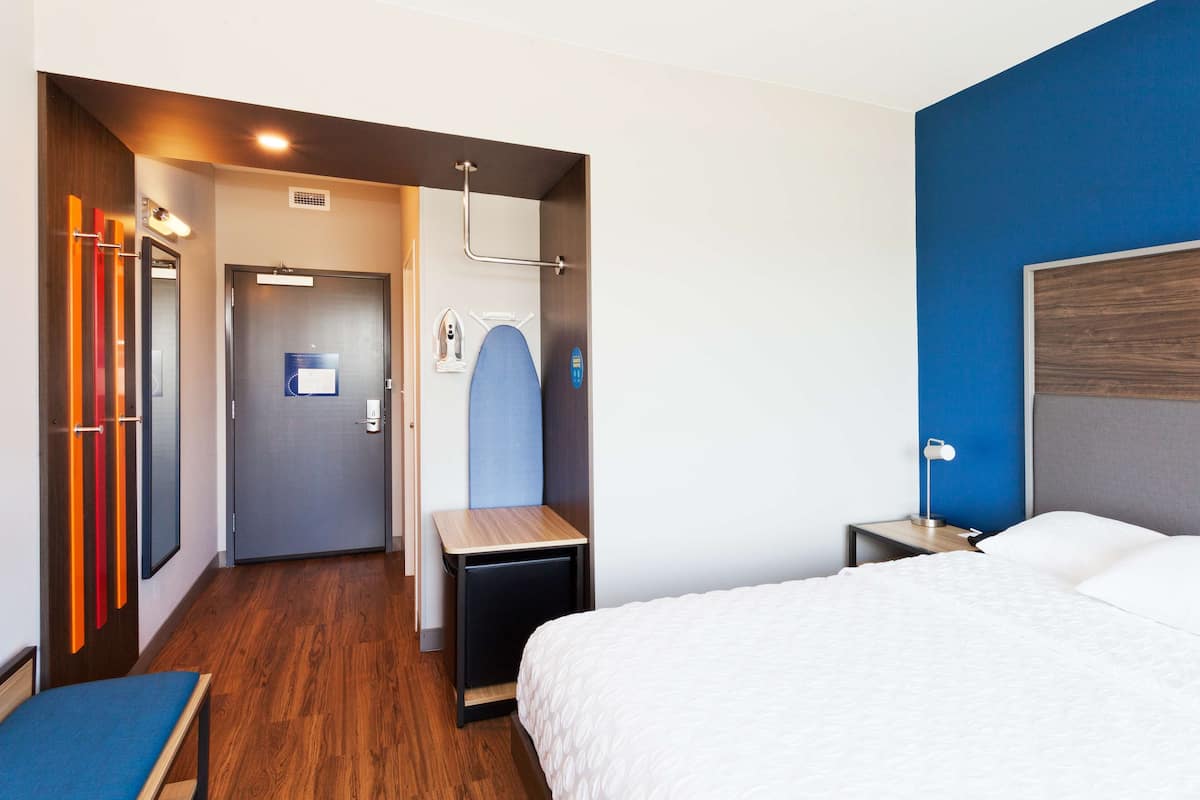 Ein Hotelzimmer mit Bett, Nachttisch und einem kleinen Schreibtisch mit Stuhl. Eine Tür führt zu einem Badezimmer und einem Flur.