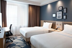 Une chambre d’hôtel avec deux lits, un bureau et une chaise. On y trouve des rideaux, un téléphone fixé au mur et des tableaux encadrés accrochés au mur.