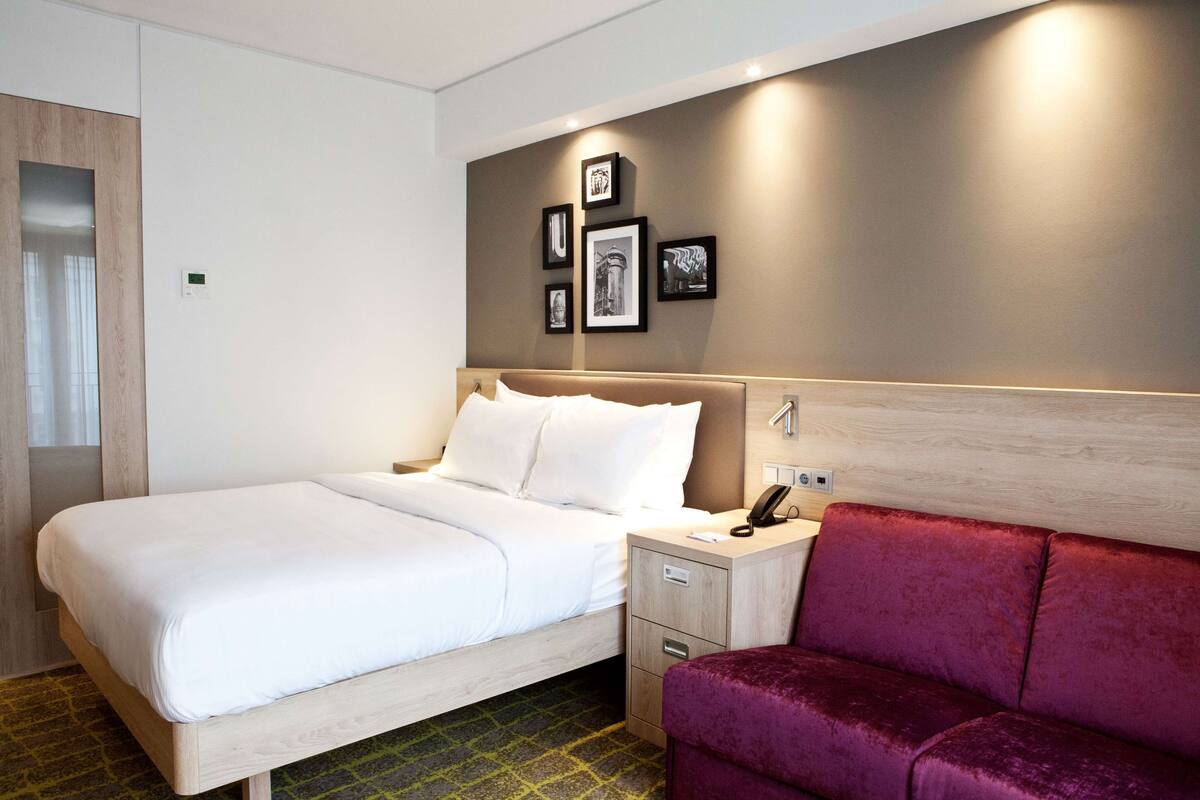 Habitación de hotel con una cama, una mesita de noche con teléfono, un sofá rojo y cuadros en la pared.