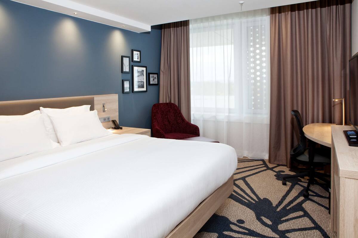 Habitación de hotel con una cama grande, un escritorio con silla, un sillón rojo y una ventana con cortinas.