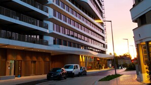 Un edificio moderno de varios pisos con fachada curva, ventanas grandes y un estacionamiento con automóviles.