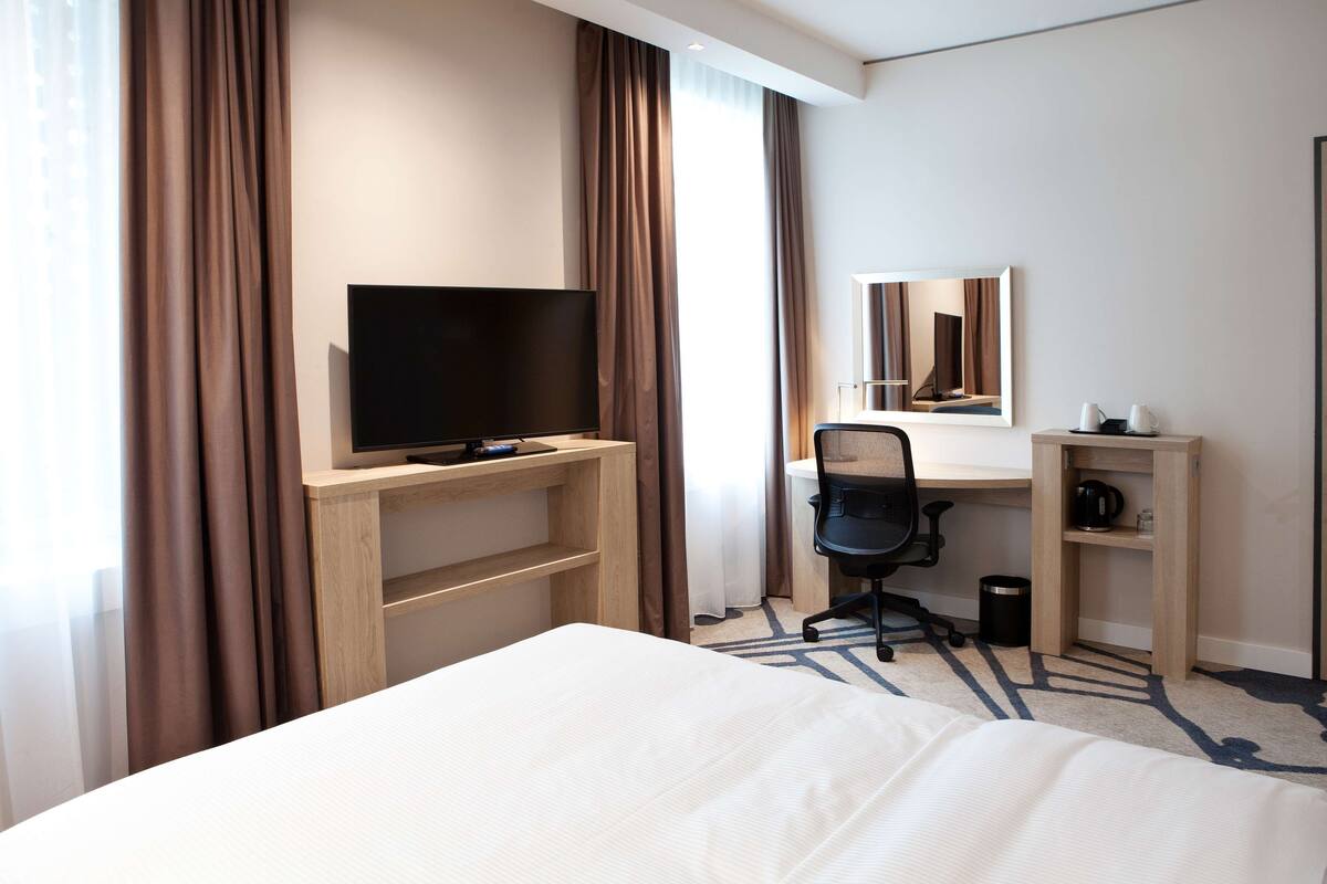 Habitación de hotel con cama, escritorio con computadora, televisor, espejo y un armario pequeño.