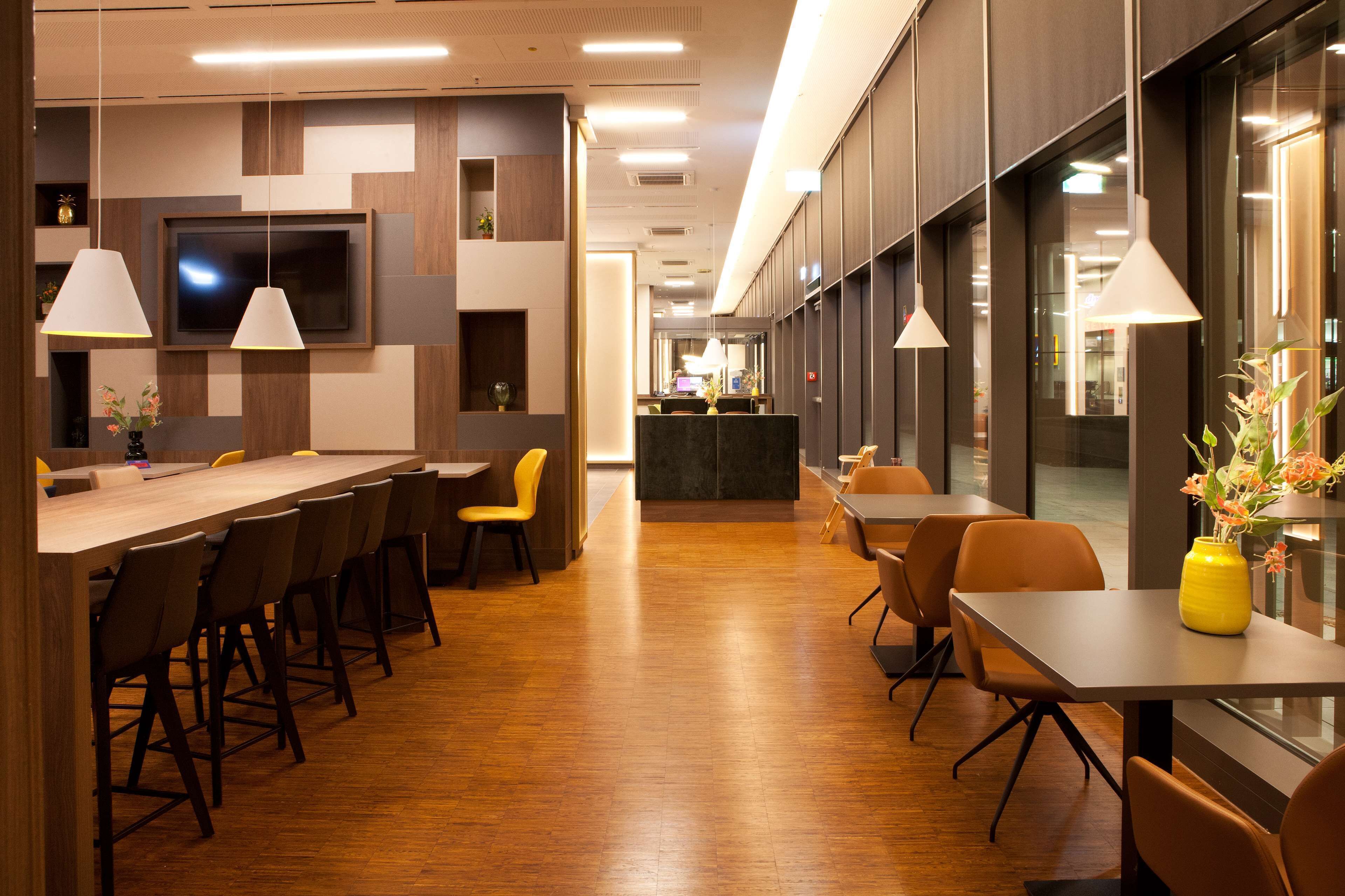 A modern hotel lobby with a long reception desk, multiple seating areas, and a large TV screen on the wall.
