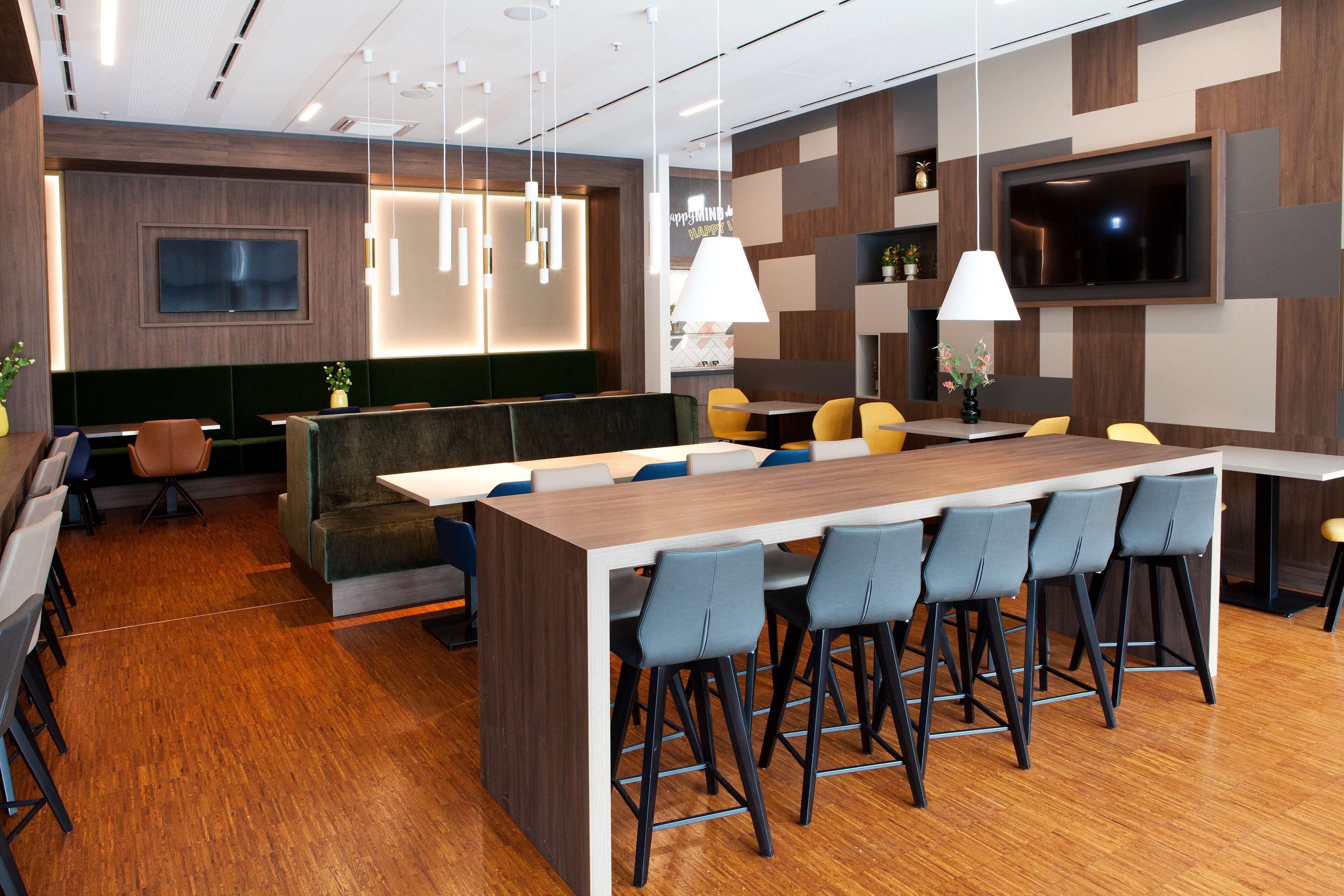 A modern restaurant with a long communal table, bar stools, and a TV mounted on the wall.
