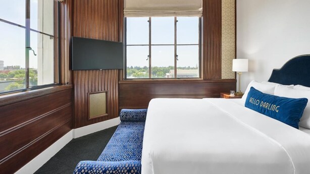 A hotel room with a large bed, a flat-screen TV mounted on the wall, a window with a view of the city, and a blue patterned sofa.