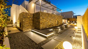 A modern outdoor patio area with stone walls, a wooden deck, and a seating area with a fire pit. - Parion Opus - Adults Only (Paros)