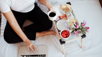 Uma pessoa sentada na cama com um laptop, um tabuleiro com comida e bebidas, e uma pequena mesa com flores.
