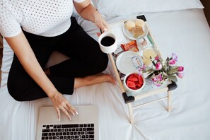 A person sitting on a bed with a laptop, a tray with food and drinks, and a small table with flowers. - Paris Hotel by H Hotéis Business (Taguatinga)