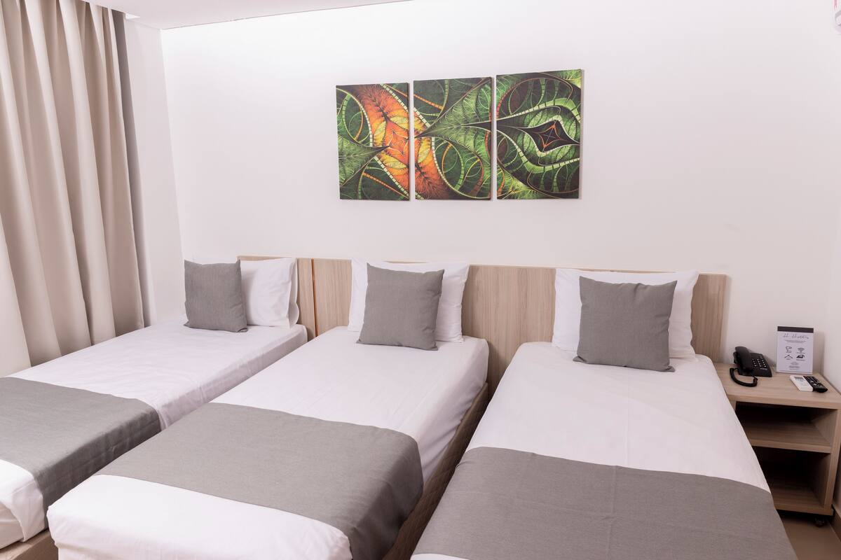 A hotel room with three single beds, each with a grey bedspread and white pillows. There is a wall-mounted artwork above the beds and a bedside table with a phone and a sign.