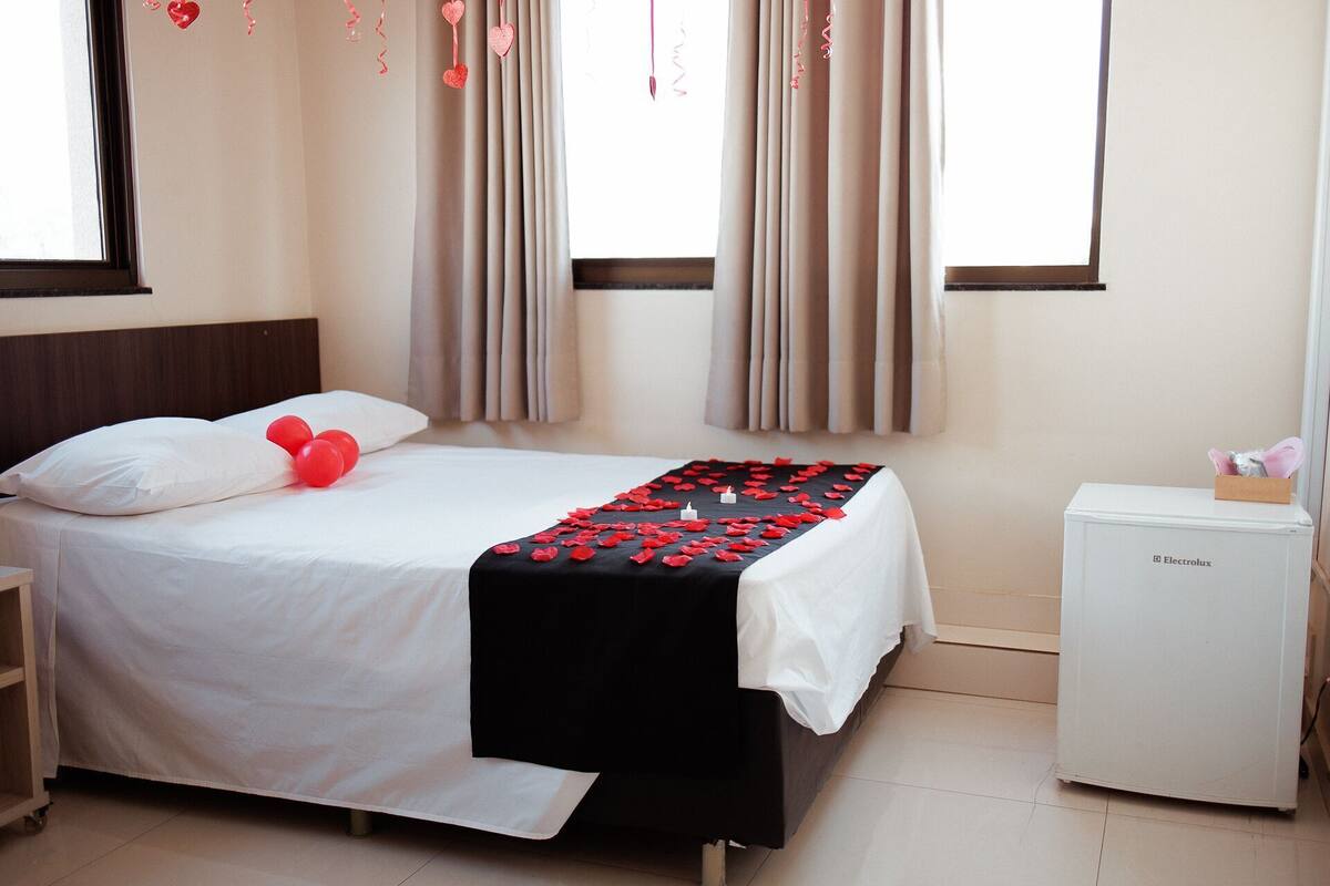 A hotel room with a bed, a nightstand, a small refrigerator, and curtains.