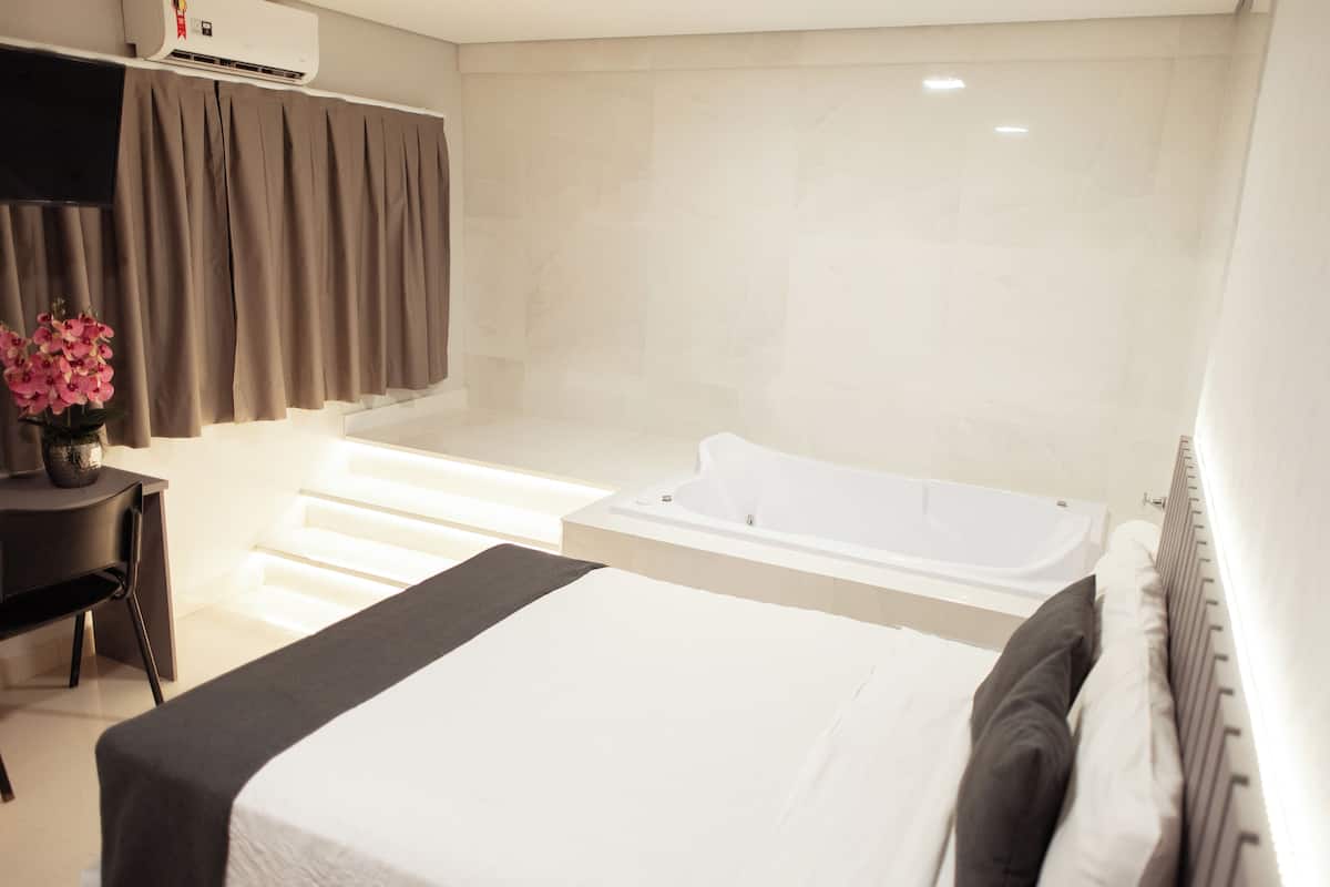 A modern hotel room with a large bed, a separate sitting area with a desk and chair, a bathtub, and a large window with curtains.