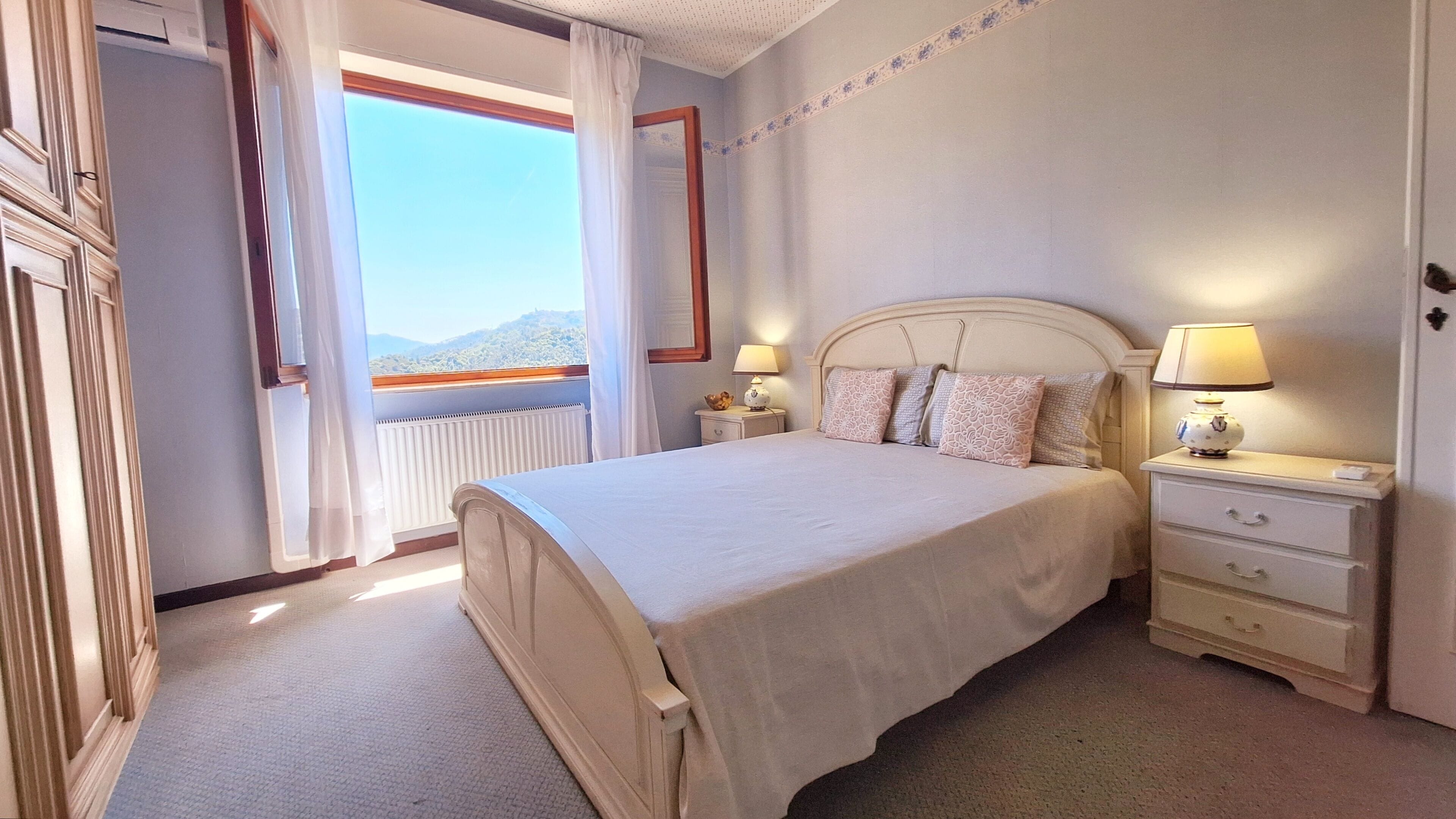 Een hotelkamer met een groot bed, twee nachtkastjes met lampen, uitzicht op de bergen door een raam en een radiator.