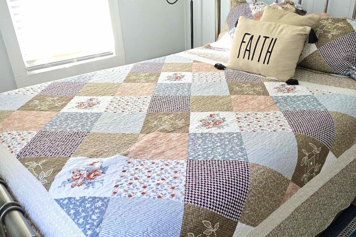 A neatly made bed with a patchwork quilt, a pillow with the word "FAITH," and a painting on the wall.