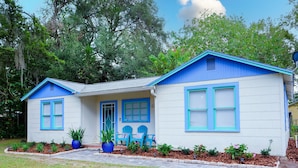 Une maison à deux étages, avec un toit bleu et des murs blancs, dotée d’un porche à l’avant, agrémenté de chaises bleues et de plantes en pot.