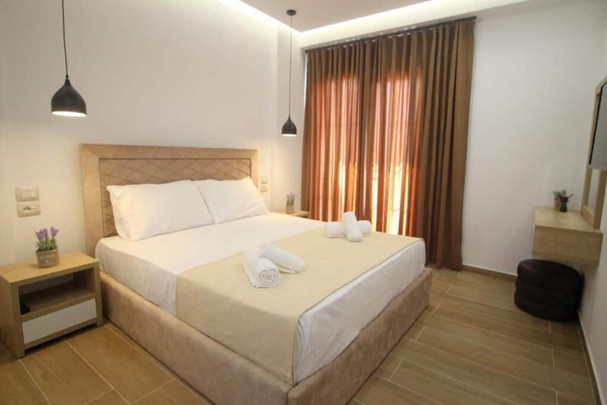 Una cama bien hecha con sábanas blancas y toallas enrolladas, una mesita de noche con una pequeña planta, y un ventanal con cortinas.