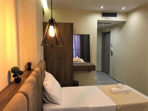 Une chambre d’hôtel avec un lit, une table de chevet ornée d’une plante, un luminaire suspendu, un petit bureau avec un miroir et une salle de bain où se trouve une serviette sur le sol.
