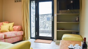 A living room with a sofa, a yellow armchair, a wooden table with cups and a bottle, and a TV on a shelf.