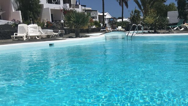 A clear blue pool with lounge chairs and palm trees. - 5 Star,  2 x True  Ensuite’s, Beach 20 meters,  Pool,  AC, FREE WIFI, Full TV.  (Puerto del Carmen)