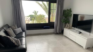 A modern living room with a black leather sofa, a white TV stand, and a large window with curtains. - 5 Star,  2 x True  Ensuite’s, Beach 20 meters,  Pool,  AC, FREE WIFI, Full TV.  (Puerto del Carmen)
