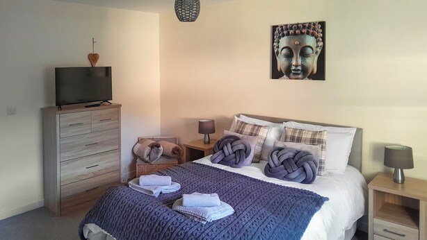 Ein aufgeräumtes Schlafzimmer mit einem Bett, einer Kommode, einem Fernseher und einem an der Wand hängenden Kunstwerk.