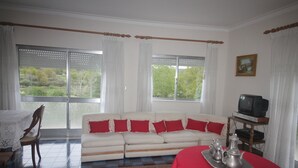Un salon avec un canapé, une table à manger recouverte d’une nappe rouge, un téléviseur posé sur un meuble en bois, et une vue sur la verdure par les fenêtres.