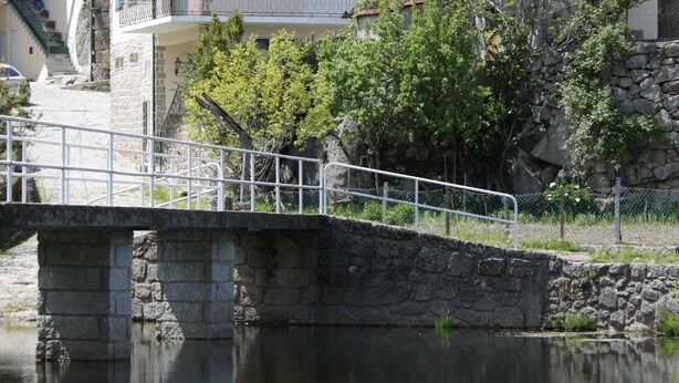 Un pont de pierre enjambe une rivière paisible, avec une zone herbeuse au premier plan et des bâtiments aux toits recouverts de tuiles rouges en arrière-plan.