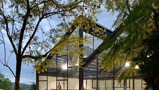 A modern glass building with a patio area, outdoor seating, and visible kitchen equipment.