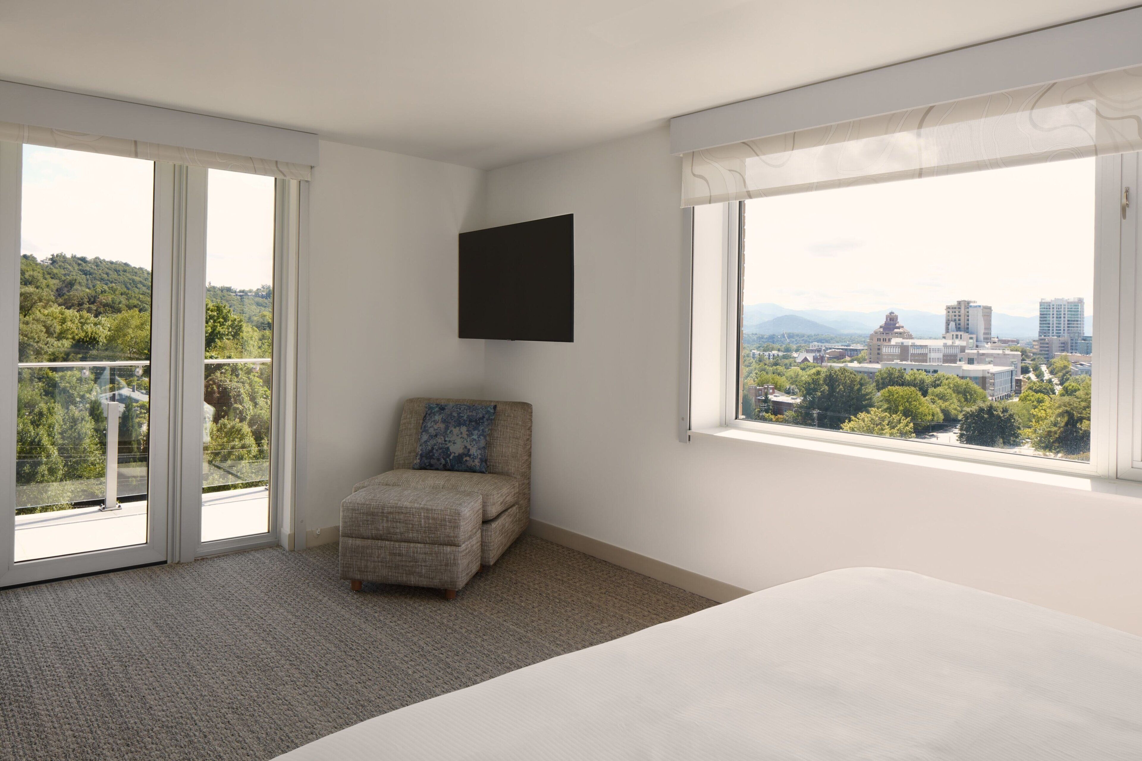 A hotel room with a large bed, a chair, a flat-screen TV, and a balcony with a city view.