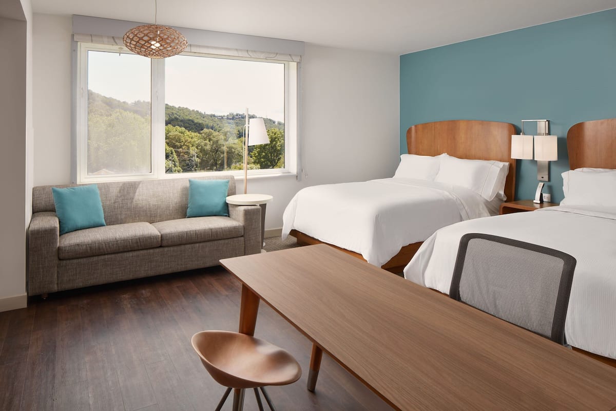 Een hotelkamer met een bank, twee bedden, een tafel en uitzicht op groen door grote ramen.