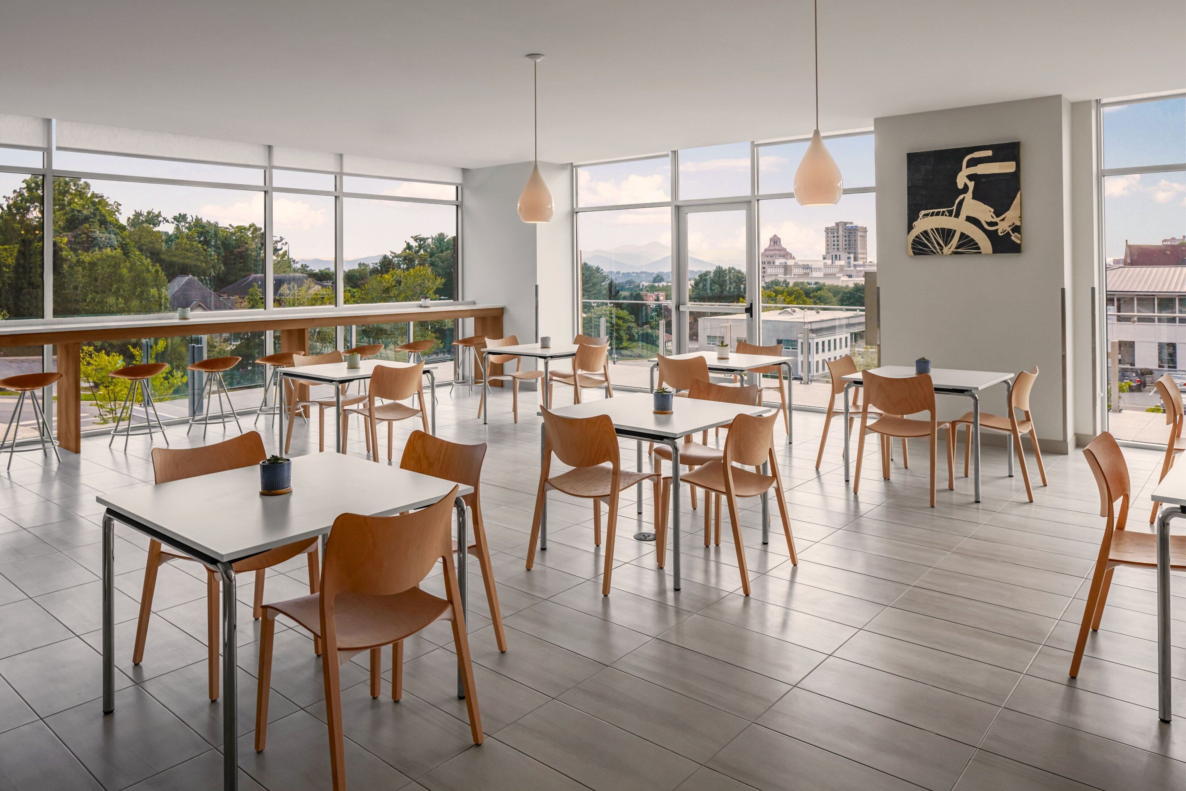 A spacious dining area with multiple tables and chairs, large windows offering a city view, and a bicycle artwork on the wall.