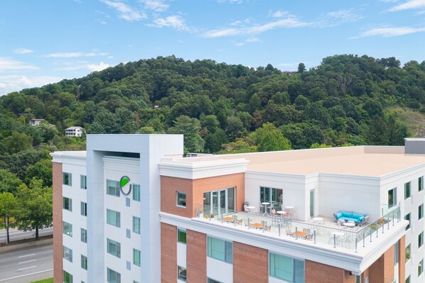 Un edificio moderno con terraza en la azotea, mobiliario exterior y vistas a una colina boscosa.