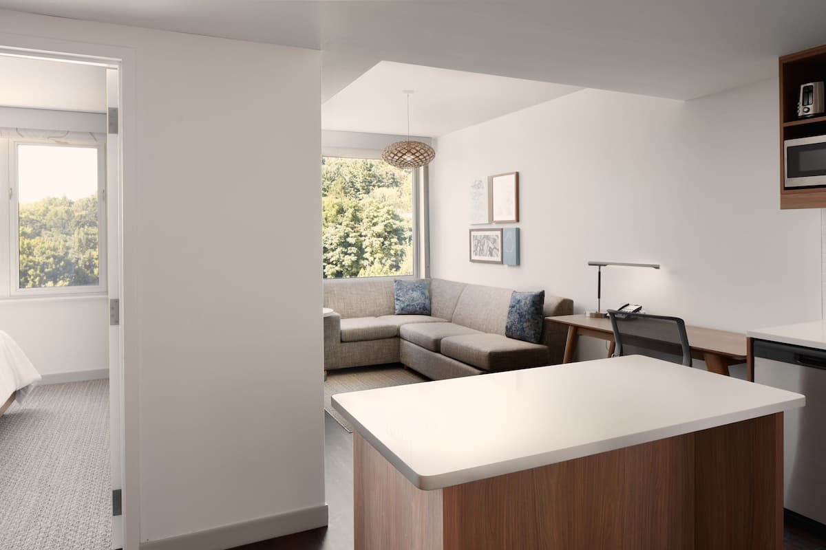 Een moderne, goed verlichte kamer met een groot raam, een bank, een eettafel en een keukenhoek.