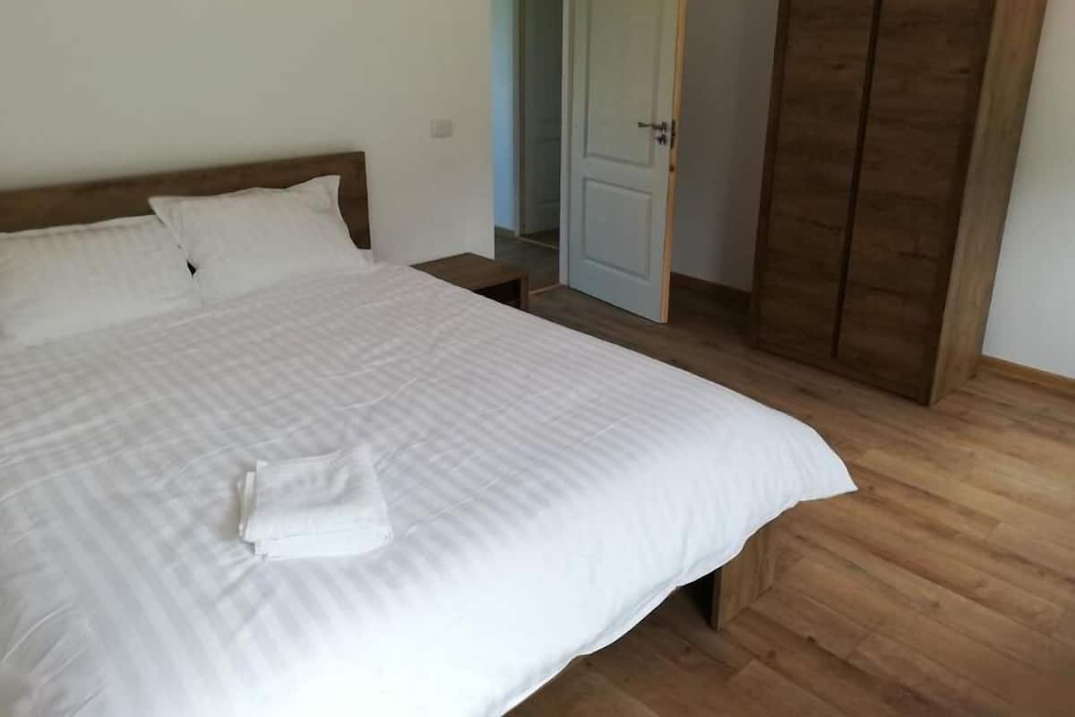 A neatly made bed with white linens, a wooden headboard, and a wooden wardrobe in a room with wooden flooring.