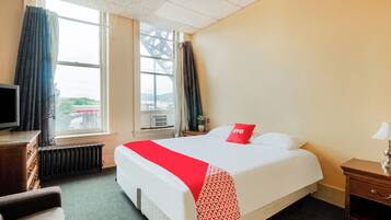 A hotel room with a bed, a TV, a window with curtains, a radiator, and a nightstand with a lamp.