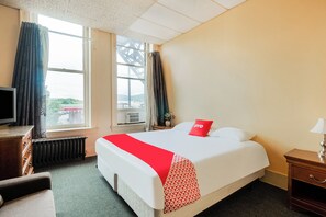 A hotel room with a bed, a TV, a window with curtains, a radiator, and a nightstand with a lamp.