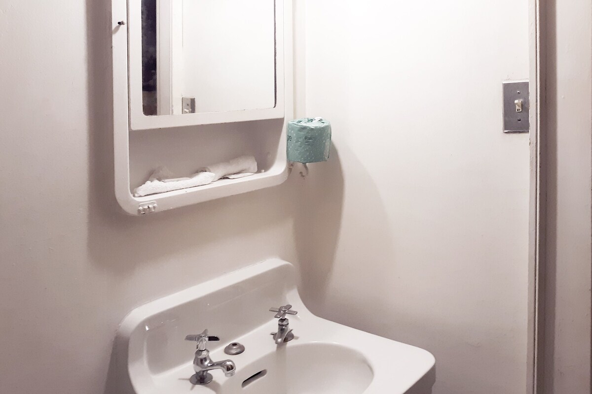 Ein Badezimmer mit Waschbecken, zwei Wasserhähnen, einem Spiegel und einem Toilettenpapierhalter.