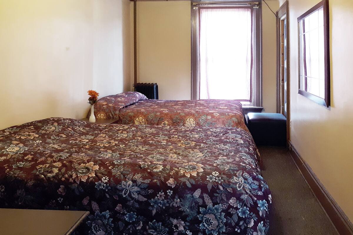 Una habitación con dos camas, una ventana con cortinas, un radiador y una mesita.