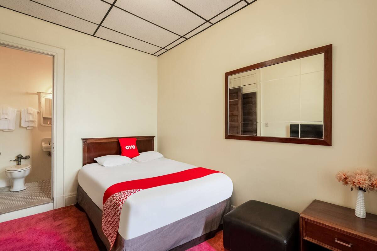 Habitación de hotel con cama, mesita de noche, espejo y baño visible a través de una puerta abierta.