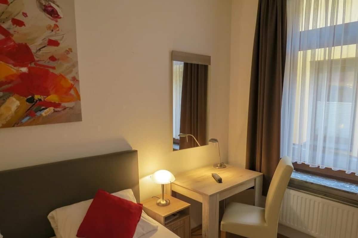 Camera d'albergo con un letto, una scrivania, una sedia, una lampada, uno specchio e una finestra con tende.